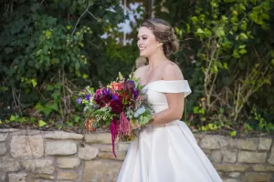Professional Photographer A smiling bride in an off-the-shoulder white gown holds a colorful bouquet while standing outdoors in front of a stone wall and leafy green foliage—perfectly captured by a talented photographer. www.maranonimagery.com