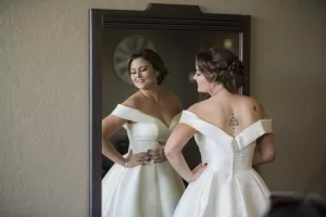 Professional Photographer A bride in a white off-the-shoulder wedding dress stands in front of a mirror, smiling at her reflection with hands on her waist. A photographer captures this candid moment, highlighting her updo and tattoo on her upper back. www.maranonimagery.com