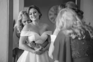 Professional Photographer A bride in an off-the-shoulder gown smiles as a woman hugs her from behind. Two others, partially visible, stand nearby. The joyful and intimate scene is beautifully captured in black and white portraits. www.maranonimagery.com