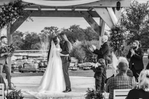 Professional Photographer A bride and groom share a kiss under a wooden gazebo during their outdoor wedding ceremony, as guests applaud and celebrate around them. This black and white scene perfectly captures the emotion of wedding photography. www.maranonimagery.com