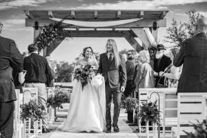 Professional Photographer A bride and a man, possibly her father, walk down the aisle outdoors under a wooden arch, smiling as wedding guests—and the photographer—capture portraits. The scene is in black and white. www.maranonimagery.com