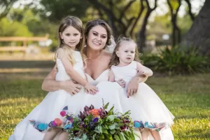 Professional Photographer A woman in a white dress kneels on grass, smiling and hugging two young girls in matching white dresses. Captured by a talented photographer, this outdoor wedding photography highlights joy against a natural backdrop and colorful bouquet. www.maranonimagery.com
