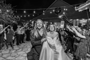 Professional Photographer A joyful bride and groom walk hand-in-hand outside at night, surrounded by cheering guests holding sparklers, under strings of lights. A talented wedding photographer captures this magical moment in front of a rustic building. www.maranonimagery.com