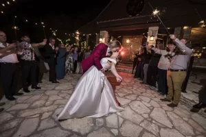 Professional Photographer A bride and groom share a romantic dip and kiss on a stone patio at night, surrounded by guests holding sparklers, as their photographer captures the magical moment outside a warmly lit building. www.maranonimagery.com