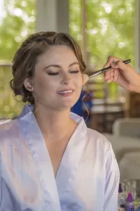 Professional Photographer A woman in a satin robe smiles with her eyes closed as someone applies makeup to her cheek with a brush—capturing the serene moment of wedding photography against a softly blurred backdrop of green foliage visible through the window. www.maranonimagery.com