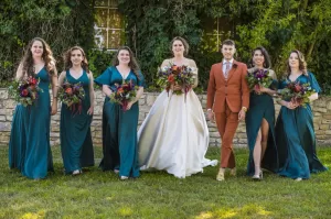 Professional Photographer A photographer captures a bride in a white gown and a groom in a rust suit, standing with five bridesmaids in teal dresses. All are holding colorful bouquets and smiling for portraits in front of a stone wall covered in greenery. www.maranonimagery.com