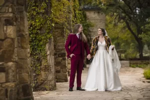 Professional Photographer A bride in a white gown and veil holds hands with a groom in a maroon suit as they walk outside on a stone path, surrounded by ivy-covered stone walls and greenery—capturing pure joy through stunning wedding photography. www.maranonimagery.com