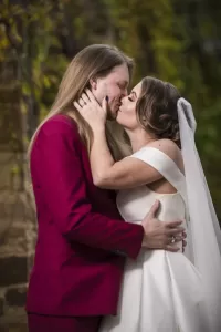 Professional Photographer A bride in a white gown and veil kisses a groom in a burgundy suit outdoors, holding each other closely with greenery in the background—a perfect moment for wedding photography portraits captured by the photographer. www.maranonimagery.com