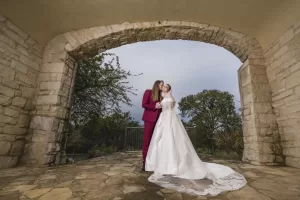 Professional Photographer A couple stands under a stone archway; one person wears a white wedding dress with a long train, the other a burgundy suit. Their photographer captures their embrace among trees and an overcast sky. www.maranonimagery.com