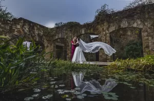 Professional Photographer A bride and groom embrace in front of stone ruins, their veil flowing dramatically in the wind. Their reflection shines in a pond surrounded by green plants—a stunning moment of wedding photography artistry. www.maranonimagery.com