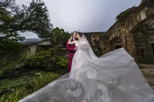 Professional Photographer A bride and groom embrace outdoors near stone ruins, with the bride’s veil flowing dramatically in the foreground—lush greenery surrounds them under a cloudy sky, captured perfectly by a skilled wedding photographer. www.maranonimagery.com