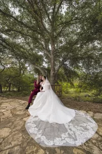 Professional Photographer A bride in a long white gown with a lace train and a groom in a burgundy suit sit close on a bench beneath a large tree, surrounded by greenery and stone pavement, sharing an intimate moment perfect for timeless portraits. www.maranonimagery.com