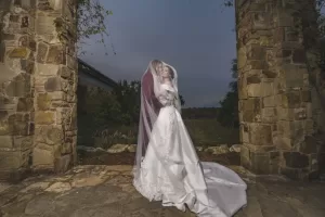 Professional Photographer A bride and groom embrace under a sheer veil between two stone pillars outdoors, captured beautifully by a wedding photographer. Surrounded by greenery and a cloudy sky, this portrait highlights her long white gown and his dark suit. www.maranonimagery.com
