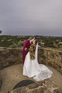 Professional Photographer A couple kisses on a stone terrace overlooking lush greenery. Captured by a talented wedding photographer, one wears a burgundy suit and the other a white wedding dress with a long veil and colorful jacket beneath an overcast sky. www.maranonimagery.com