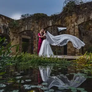 Professional Photographer A couple in wedding attire embraces in front of stone ruins near a pond; the bride’s long veil flows dramatically in the wind, capturing a timeless moment of wedding photography reflected beautifully in the water below. www.maranonimagery.com