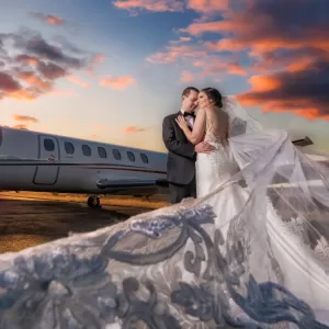 Professional Photographer A bride and groom embrace in front of a private jet at sunset. The bride’s long, flowing veil billows dramatically, making this moment truly worthy of luxury portraits against a sky filled with colorful clouds. www.maranonimagery.com