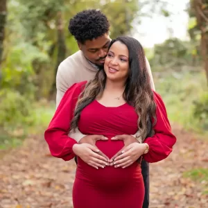 Professional Photographer A man embraces a pregnant woman from behind, placing his hands over her belly. The woman in a red dress smiles, while the man looks at her lovingly. This heartwarming moment is perfect for family portraits on a leafy path surrounded by greenery. www.maranonimagery.com
