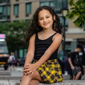 Professional Photographer A young girl with long curly hair, wearing a black tank top and a yellow plaid skirt, sits on a stone ledge outdoors. Captured by a talented photographer, she smiles with her hands on her knee, framed against a blurred cityscape backdrop. www.maranonimagery.com