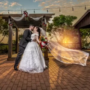 Professional Photographer A photographer captures a bride and groom kissing outdoors at sunset under a decorated wooden pergola; the bride’s long veil flows dramatically behind her as she holds a colorful bouquet, with string lights glowing above them. www.maranonimagery.com