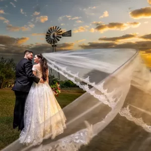 Professional Photographer A bride and groom stand outdoors at sunset near a windmill. The bride’s long veil flows dramatically in the wind as they gaze at each other lovingly, creating a portrait-worthy moment against the colorful sky and clouds. www.maranonimagery.com
