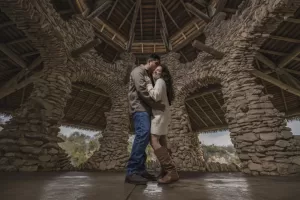 Professional Photographer A couple stands closely together, embracing under a rustic stone gazebo with arched openings and a wooden roof. Captured by a skilled photographer, natural light fills the structure as they share this intimate moment. www.maranonimagery.com
