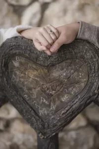 Professional Photographer Two hands gently hold each other atop a wooden heart-shaped carving marked with various etched initials; one hand wears an engagement ring with a turquoise stone—a perfect moment for any photographer to capture. www.maranonimagery.com