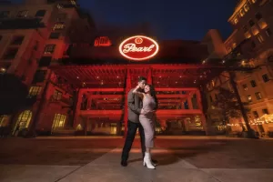 Professional Photographer A couple poses together at night in front of the illuminated Pearl sign in an urban plaza, as a photographer captures their moment amid historic brick buildings and warm lights. www.maranonimagery.com
