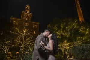 Professional Photographer A couple shares a kiss at night in front of a lit-up historic building and twinkling trees, surrounded by greenery and festive lights—capturing the romance of wedding photography in a magical setting. www.maranonimagery.com