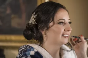 Professional Photographer A woman with dark hair styled in an elegant updo and adorned with a jeweled hair accessory smiles as someone applies makeup to her lips with a brush—a beautiful moment captured in wedding photography. She is wearing a cozy blue and white patterned robe. www.maranonimagery.com