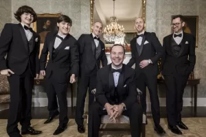 Professional Photographer Six men in black tuxedos pose indoors for elegant portraits. Five stand behind a seated man, all smiling. The photographer captures them in a refined room with a chandelier, ornate mirror, classic wallpaper, and framed artwork. www.maranonimagery.com