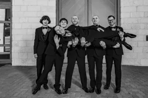 Professional Photographer Five men in tuxedos pose for a classic wedding photography portrait; four are standing and smiling while holding up the fifth man horizontally in front of a stone building. The black and white image captures a memorable moment. www.maranonimagery.com