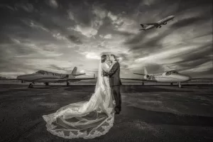 Professional Photographer A photographer captures a bride and groom kissing on an airport runway between two private jets, with a commercial airplane soaring overhead in a dramatic, cloudy sky. The black-and-white portrait features the bride’s veil flowing elegantly on the ground. www.maranonimagery.com
