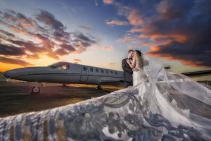 Professional Photographer A bride and groom embrace beside a private jet at sunset, with dramatic clouds overhead and the bride’s lace veil billowing toward the camera—a stunning example of wedding photography. www.maranonimagery.com