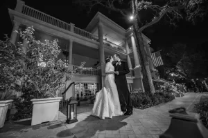 Professional Photographer A bride and groom stand close together at night outside a large, elegant building with balconies and columns, surrounded by trees and potted plants—a romantic black-and-white wedding photography portrait. www.maranonimagery.com