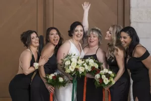 Professional Photographer A bride in a white dress and five bridesmaids in black dresses stand together, smiling and laughing, holding bouquets of flowers in front of wooden doors—a joyful moment captured through wedding photography. www.maranonimagery.com