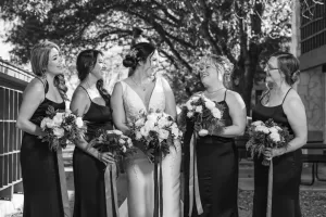 Professional Photographer A photographer captures a bride in a white dress outdoors with four bridesmaids in matching dark gowns, all holding bouquets and smiling at each other under trees in bright sunlight. www.maranonimagery.com