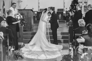 Professional Photographer A photographer captures the bride and groom kissing at the altar during their wedding ceremony in a church, as guests applaud. The bride’s long veil and train add elegance to a scene filled with flowers and candles. www.maranonimagery.com