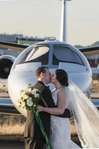 Professional Photographer A bride and groom share a kiss in front of a small private jet. The bride, holding a bouquet and wearing a long veil, stands with the groom in his suit—an elegant moment captured through wedding photography at an outdoor airport setting. www.maranonimagery.com
