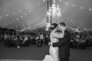 Professional Photographer A bride and groom share their first dance under a tent with string lights and chandeliers, captured beautifully in wedding photography as guests seated at tables watch and smile in the background. www.maranonimagery.com