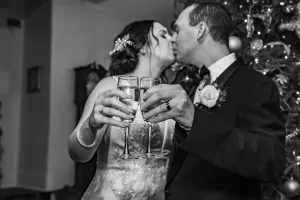 Professional Photographer A bride and groom share a kiss while holding champagne glasses in a toast, standing before a decorated Christmas tree. This black and white portrait beautifully captures the magic of wedding photography. www.maranonimagery.com