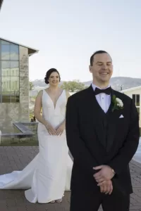 Professional Photographer A bride in a white gown stands smiling behind a groom in a black tuxedo with a white boutonnière, who is smiling with his eyes closed, outdoors near modern buildings—capturing the joy of wedding photography. www.maranonimagery.com