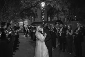 Professional Photographer A bride and groom kiss under a streetlight at night, surrounded by guests holding candles and forming two lines. The scene is festive and elegant, exemplifying timeless wedding photography in exquisite black and white. www.maranonimagery.com