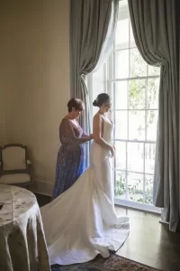 Professional Photographer A woman in a sparkling dress helps a bride in a white gown adjust her dress near a tall window with curtains, sunlight streaming in and greenery outside—a perfect moment for wedding photography. www.maranonimagery.com