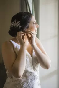 Professional Photographer A bride in a white wedding dress smiles as she stands by a window, putting on an earring. Her dark hair is styled in an updo with a decorative hairpiece, and sunlight softly illuminates her face as the photographer captures the moment. www.maranonimagery.com