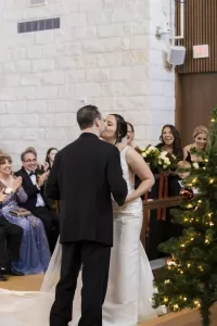 Professional Photographer A bride and groom share a kiss at the altar during their wedding ceremony, while guests behind them smile and cheer. A decorated Christmas tree is visible in the foreground, capturing a magical moment of wedding photography. www.maranonimagery.com