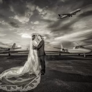 Professional Photographer A bride and groom kiss on an airport runway, surrounded by private jets, with a commercial airplane flying overhead against a dramatic, cloudy sky in this striking black-and-white wedding photography portrait. www.maranonimagery.com