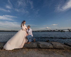 Professional Photographer A man kneels and kisses the belly of a pregnant woman in a flowing dress, both by the water with a bridge and blue sky in the background—a tender moment perfect for wedding photography portraits. www.maranonimagery.com