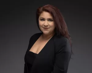 Professional Photographer A woman with long, dark red hair wearing a black blazer and top poses against a dark grey background, smiling softly at the camera—an elegant example of modern headshots. www.maranonimagery.com