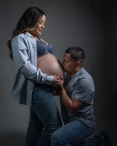 Professional Photographer A man kneels and kisses the bare belly of a pregnant woman who is smiling and gently holding his hand. The scene evokes the warmth often found in intimate portraits, with dim lighting and a neutral background adding to the mood. www.maranonimagery.com
