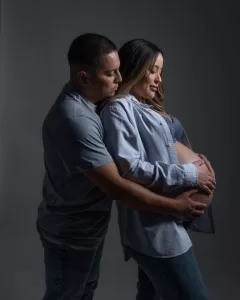 Professional Photographer A photographer captures a tender moment as a man stands behind a pregnant woman, gently holding her belly. Both in casual blue clothing, they pose together in a studio setting with a dark background. www.maranonimagery.com